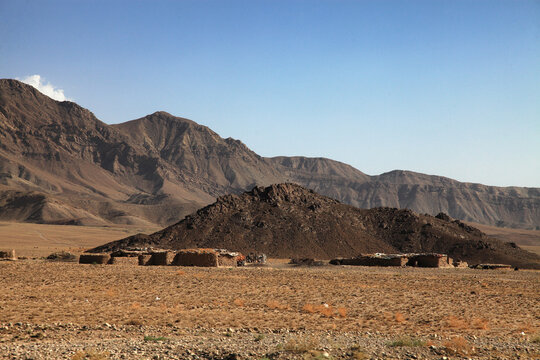 View of rugged, arid landscape with simple dwellings nestled against a dark, rocky outcrop under the gaze of towering mountains, Quetta, Balochistan, Pakistan.