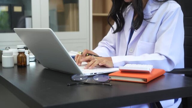 Asian female doctor using laptop for teleconsultation and checking patient's EHR record, wearing glasses,demonstrating modern healthcare,online medical services  professional expertise in hospital