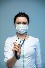 Female medical worker in mask showing stethoscope toward camera while wearing white uniform