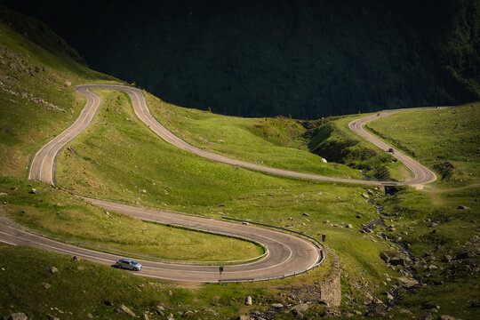 View of a winding asphalt road snakes through the vibrant green hillsides, bathed in sunlight, carving a path through the rugged landscape, Poienari, Transilvania, Romania.