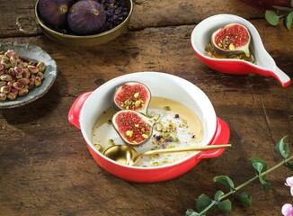 Halved fresh figs are presented in a creamy sauce, adorned with pistachios and served in red and white bowls. A golden spoon rests nearby, promising a sweet indulgence