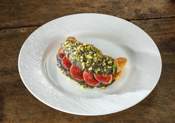 A flaky croissant, adorned with rich chocolate, sweet fig slices, and crushed pistachios, sits elegantly on a textured white plate, ready to be enjoyed