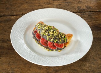 A golden croissant rests on a white plate. It is filled with slices of fresh figs, drizzled with dark chocolate, and sprinkled with chopped pistachios