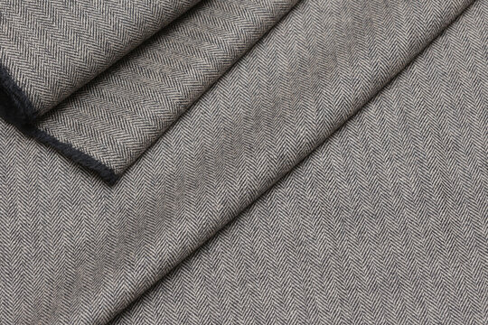 Close-up view of classic English tweed wool fabric in a fine gray and black herringbone weave. Detailed texture and folded pattern perfect for tailoring suits, coats, and blazers.