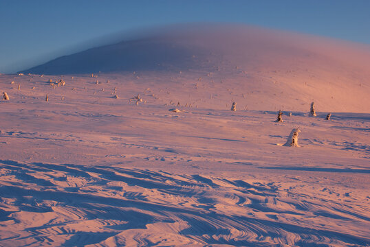 View of a serene, snow-covered landscape kissed by the warm light of dusk, with windswept patterns and a cloud-veiled mountain, Velka Hola, Banska Bystrica Region, Slovakia.