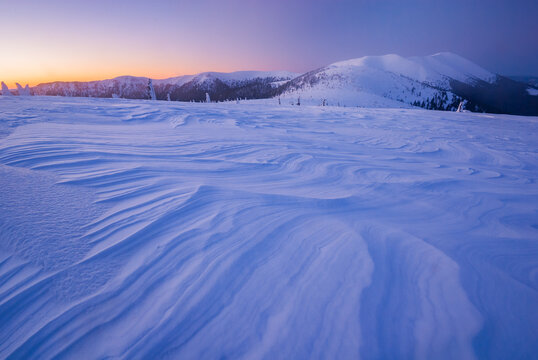 View of wind-sculpted snowdrifts in the foreground lead the eye to snow-covered mountains under a pastel sunrise sky, Velka Hola, Banska Bystrica Region, Slovakia.