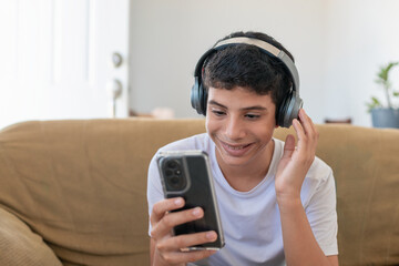 A young man is sitting on a couch and looking at his cell phone. He is wearing headphones and he is listening to music © Dionisio