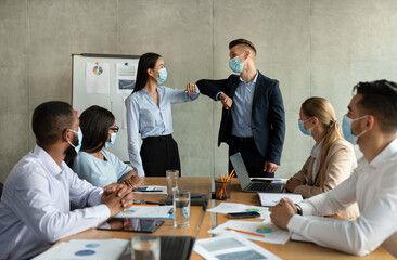 Two coworkers in medical masks are greeting each other with elbow bumps in a modern office during a corporate meeting. A group of professionals is working collaboratively amidst the pandemic.