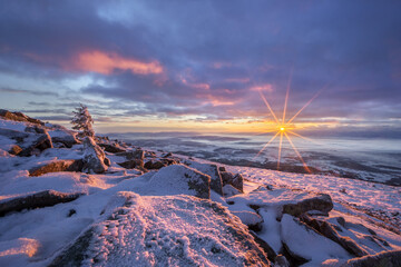 View of a serene, snow-covered landscape where the sun bursts through the horizon, casting a warm glow on the icy rocks, Babia Hora, Zilina Region, Slovakia.