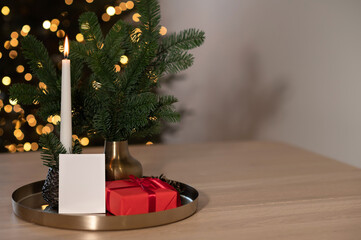 Festive Christmas holiday celebration backdrop, red gift box, candle light and blank paper card mockup on table with empty copy space, blurred Christmas tree lights bokeh on background