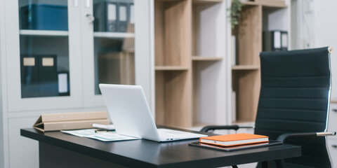 Fototapeta premium A modern office workspace with a black desk, documents, a coffee cup, and a laptop. The empty ergonomic chair and neat setup reflect professionalism, organization, and corporate productivity