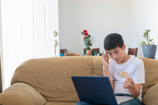 A boy is sitting on a couch with a laptop in front of him. He is holding a credit card and looking at it. The scene suggests that he might be checking his bank account or making a purchase online