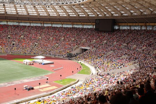 A stadium full of people watching a race. The crowd is packed in the stands, and there are umbrellas being held by some of the spectators