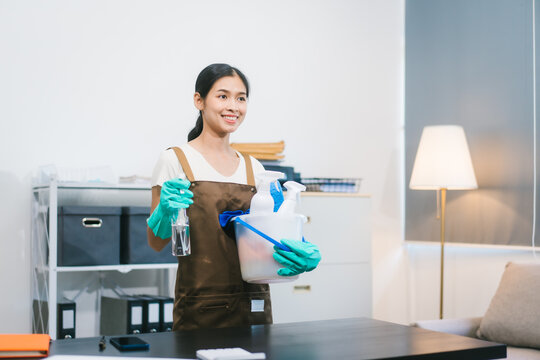 An Asian woman cleaner wearing gloves is wiping a computer monitor with a cloth in a modern office. She works carefully and smiles, maintaining cleanliness and hygiene in the workspace - Powered by Adobe