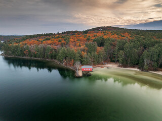 Aerial view of a red boathouse nestled at the lake's edge, surrounded by autumn foliage under a moody sky, Alton, New Hampshire, United States.