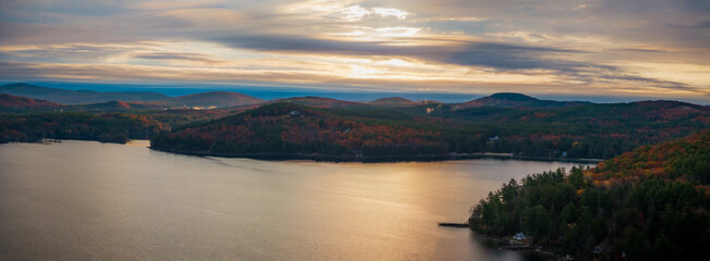 Aerial view of the lake's glassy surface reflects the fiery sunset, framed by autumn-tinged forests and distant, hazy mountains, Alton, New Hampshire, United States.