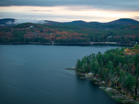 Aerial view of the tranquil lake mirroring the sky, framed by the vibrant autumn foliage, with houses nestled along the shoreline, Alton, New Hampshire, United States.