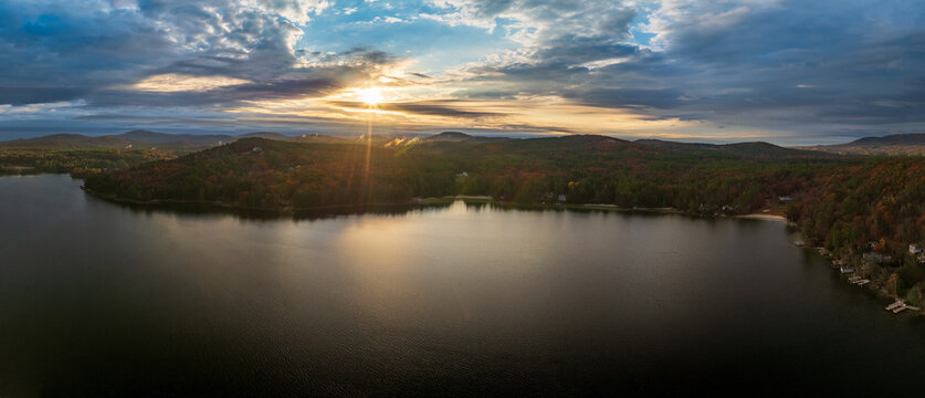 Aerial view of the sun's golden rays piercing through the clouds and reflecting on the tranquil waters of the lake surrounded by forests, Alton, New Hampshire, United States.