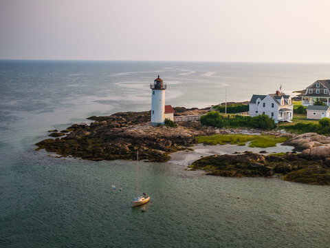 Aerial view of the iconic Eastern Point Lighthouse standing tall against the serene ocean, with a lone sailboat gently bobbing in the turquoise water, Gloucester, Massachusetts, United States.