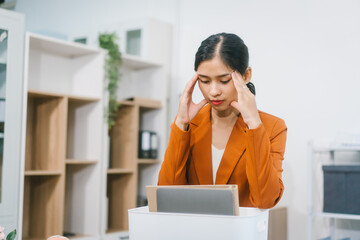 A young Asian woman in a formal blazer packs her belongings into a box while holding a tablet,...