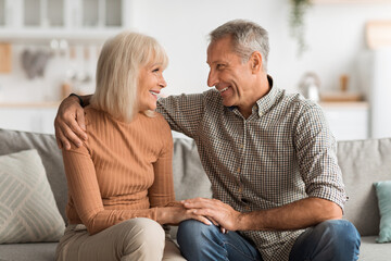 Two older adults enjoy a joyful moment together on a sofa at home. They smile and engage in conversation, creating a warm and loving atmosphere in their living room.