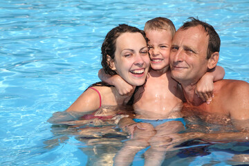 A family of three is in the water, with a woman and a child hugging a man. Scene is happy and joyful, as the family senior is enjoying their time together in the pool