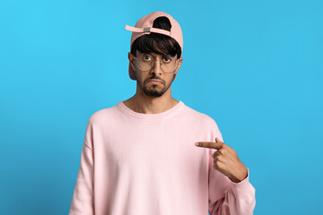 A young man wearing a pink sweater and cap poses against a bright blue background. He points at himself with a playful expression, expressing fun and confidence in a casual setting.