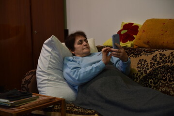 Elderly woman lying on a couch under a blanket, propped up by pillows while using a smartphone © Oleksandr