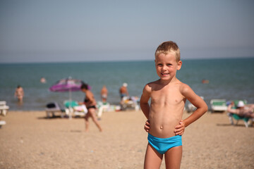A young boy stands on a beach with his arms crossed and a smile on his face. The beach is crowded with people, some of whom are standing in the water