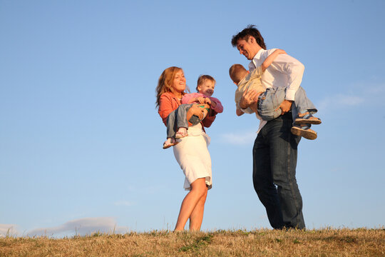 A family of four is playing together on a grassy hill. The man is holding two children while the woman is holding another child. The family is enjoying their time together in the outdoors - Powered by Adobe