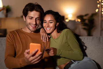 Couple sits on their couch at home, happily exploring a new application on their cellphone. The wife excitedly points at the screen, sharing great app suggestions with her spouse.
