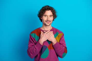 Young man with colorful sweater stands against blue background smiling with hands over heart portrait