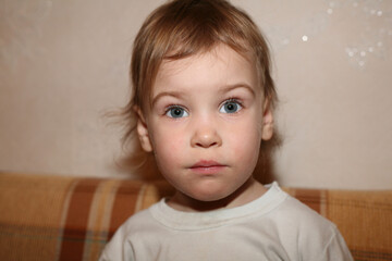 Fototapeta premium A young child with blonde hair and blue eyes is sitting on a couch. The child is wearing a white shirt and he is looking at the camera