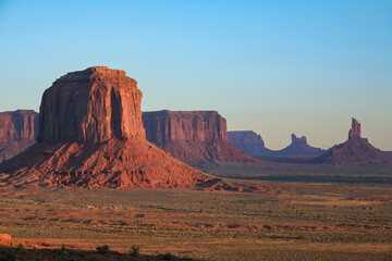 Sunrise over Monument Valley with iconic sandstone buttes glowing in warm light, casting long shadows across the vast desert landscape of the American Southwest.