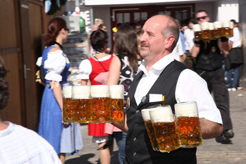 A man wearing a vest is holding a tray of beer glasses. The man is surrounded by people, some of whom are wearing traditional clothing. The atmosphere appears to be festive and lively
