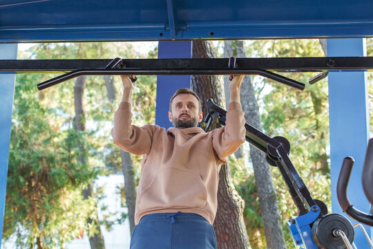 Man in a beige hoodie doing pull-ups on an outdoor bar in a forest park, training strength and upper body muscles in natural daylight, showcasing active lifestyle, fitness routine and healthy habits - Powered by Adobe