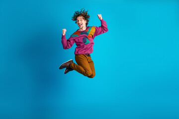 Fototapeta premium Young man jumps in the air wearing a colorful sweater against a blue background for a lively fashion lifestyle image
