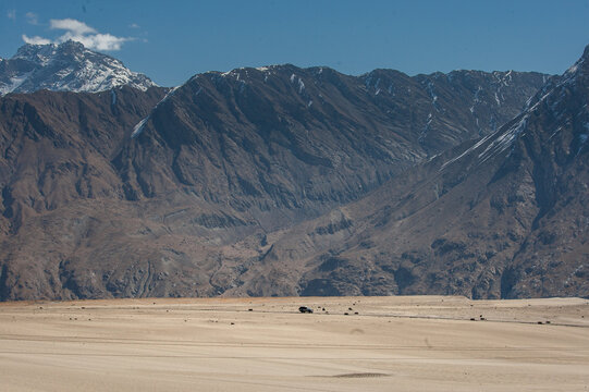 View of rugged, arid mountains rise majestically behind a vast, sandy plain under a clear blue sky, creating a stark contrast of textures and colors, Shigar, Gilgit Baltistan, Pakistan.