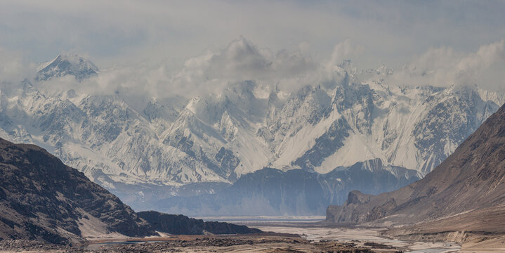 View of stark, rocky terrain gives way to majestic snow-capped mountains piercing the sky, partially veiled in soft, ethereal clouds, Shigar, Gilgit Baltistan, Pakistan.
