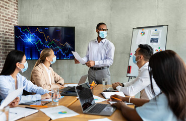 A black CEO in a face mask discusses company achievements with colleagues during a business meeting in a modern office setting. The team is engaged in the presentation with charts displayed.