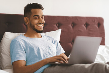 A young man sits comfortably in bed, smiling as he interacts with his laptop in a cozy room. The soft lighting creates a relaxed atmosphere while he enjoys his morning.