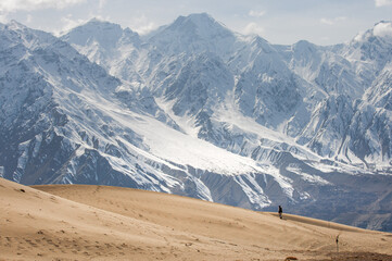 View of a lone figure treks across undulating golden sand dunes contrasted against the majestic, snow-capped peaks rising in the distance, Skardu, Gilgit Baltistan, Pakistan.