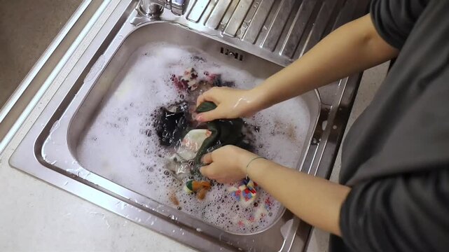 Female hands scrubbing colorful patterned socks under foam water inside metal sink, bright fabric tones visible through soapy surface, manual laundry action showing grip, splash, texture contrast, wet