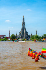 View of Wat Arun across the Chao Phraya River in Bangkok, Thailand, captured on a bright sunny day with the temple iconic spires rising above the shimmering water.