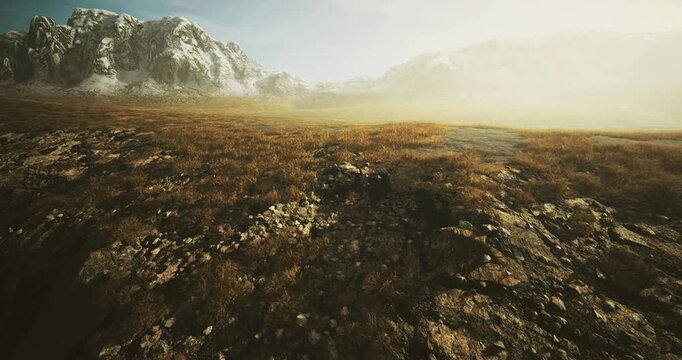 A vast expanse of rocky terrain covered with sparse grass is illuminated by soft sunlight. Surrounding mountains loom in the background, creating a serene and tranquil atmosphere.