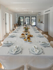 An elegant festive dining hall is prepared for guests, with a long white table set with plates, glasses and napkins, while assorted appetizers are arranged along the center ready to share in a meal