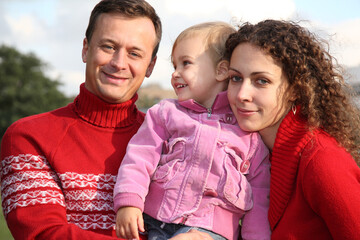 A family of three, a man, a woman and a child, are posing for a picture. The man is wearing a red sweater and the woman and child are wearing pink. Scene is warm and happy