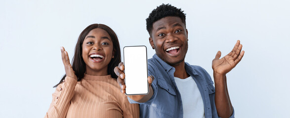 A young couple stands together, both smiling widely. They are holding a blank smartphone in front of them. The background is light and simple, adding to the joyful mood.