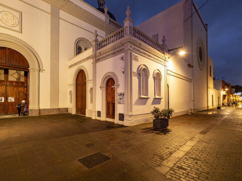 San Cristobal de La Laguna, Spain - 17 December 2016: View of a pristine white building with elegant arches and a warmly lit doorway, casting a soft glow onto the dark, glistening cobblestone street.