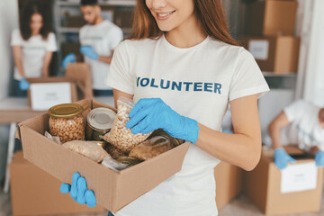 A group of volunteers sorts food items at a community center. A woman holds a box filled with various canned goods and dried food while wearing blue gloves.